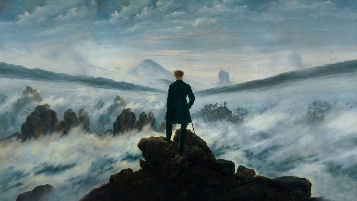 Man in a dark coat with a walking stick stands on a rocky summit above swirling fog, with distant peaks and cloudy sky