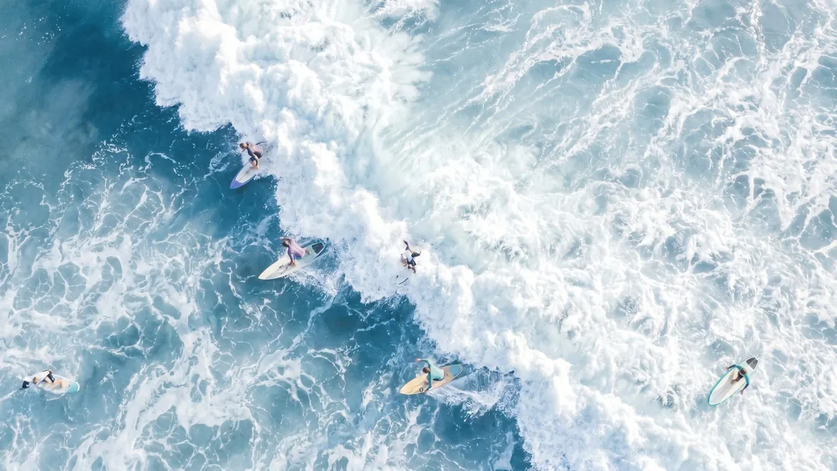 Aerial view of surfers on boards among white foamy waves breaking across deep blue water